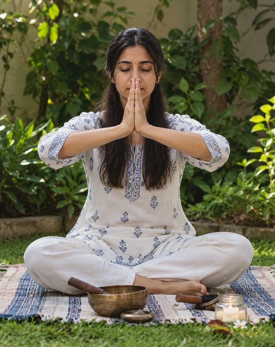Woman seated cross-legged in a garden with hands in prayer position, a singing bowl and candle in front of her